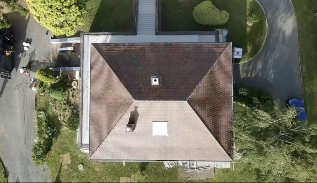 Vue aérienne d'une toiture en tuiles brunes encrassées, avec souche de cheminée et fenêtre de toit